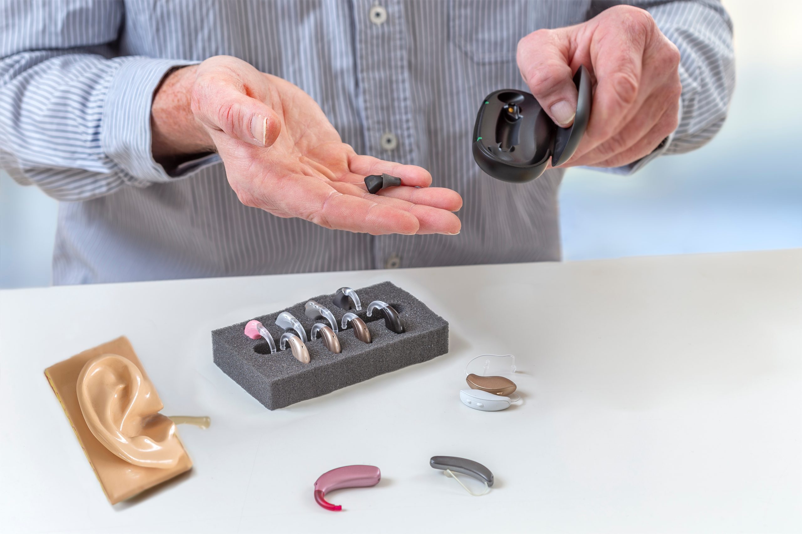 A person examines various hearing aids laid out on a table. They hold one device in their hand, reviewing its design while other aids remain in a foam tray nearby.
